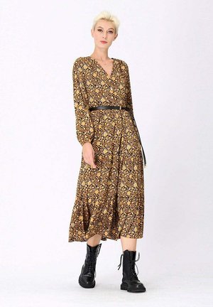 Robe à manches longues, longueur genou en moutarde foncé avec imprimé floral. Elle dispose d'un décolleté en V, d'une taille ceinturée et d'une jupe évasée. Associée à des bottes de combat noires.