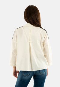 Camisa ligera, color crema, con un pliegue trasero fruncido, acentos negros en los hombros y mangas anchas y distintivas. Combinada con jeans azules.