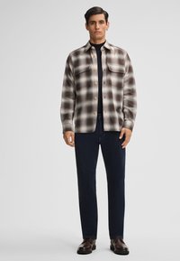 Camicia a scacchi in crema, marrone e nero; con bottoni e due tasche sul petto. Indossata con pantaloni blu scuro e scarpe marroni.