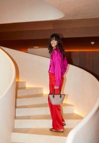Helles pinke Satinbluse und rote Hosen, eine gewebte schwarz-weiße Tasche tragend, eine kurvenreiche Treppe mit warmem Licht hinaufsteigend.