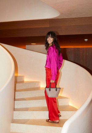 Bright pink satin blouse and red trousers, carrying a woven black and white bag, ascending a curved staircase with warm lighting.