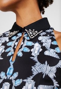 Robe en tissu noir avec un col orné de strass clairs et bleus, des motifs complexes et une ouverture en forme de trou de serrure.