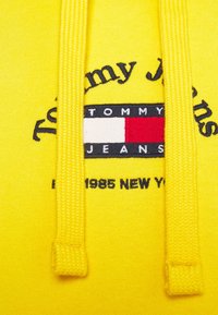 Sweat à capuche jaune avec texte "Tommy Jeans" brodé en noir, logo Tommy Hilfiger rouge, blanc et noir, et cordons jaunes à l'avant.