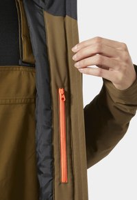 Main tenant une veste marron ouverte, montrant une poche intérieure verticale à hauteur de la poitrine avec une fermeture éclair orange fermée, sur une doublure noire.