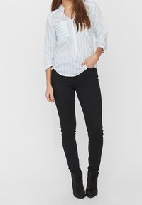 Žena oblečená v bílo-modré pruhované blůze s dlouhým rukávem, černých skinny kalhotách a černých kotníkových botách, stojící s jednou rukou v kapse.