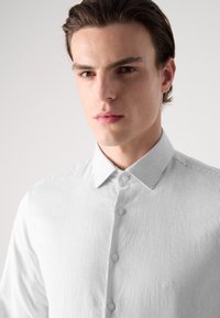 Weiß gestreiftes Hemd mit Button-Down-Kragen. Verfügt über kleine Knöpfe und ein dezentes Logo auf der linken Brust. Glatte Textur.