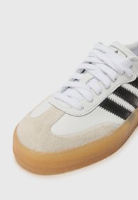 Zapatilla blanca con franjas negras, materiales de ante y cuero, puntera redonda, cordones blancos, suela de goma con textura.
