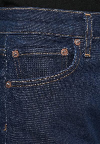 Tumed sinised teksapüksid vaskse tooniga riistvaraga, mis on viie taskuga ja kontrastseks oranžiks tikkimisega. Sile tekstuur ja õrn läige.