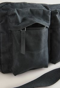 Sort stofpose med en tekstureret overflade, der har en lynlåslukket frontlomme og en bred, justerbar strop. Enkel rektangulær form.