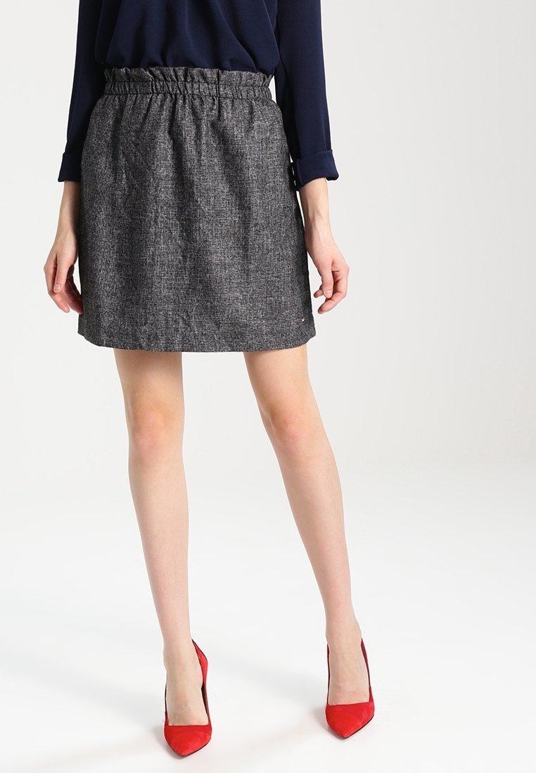 Žena v šedé texturované sukni, námořnickém modrém topu a červených vysokých podpatcích stojí na bílém pozadí.