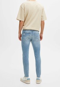 Ljusbeige t-shirt med korta ärmar och avslappnad passform, tillsammans med ljusblå slim-fit jeans med slitna detaljer och sneakers med blå accenter.