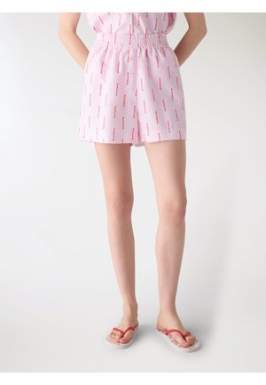 Donna con pantaloncini rosa stampati e infradito bianchi con cinturini rossi, in piedi con le mani dietro la schiena su sfondo bianco uniforme.