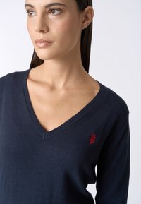 Maglione a V navy realizzato in tessuto morbido, con maniche lunghe e un piccolo logo ricamato rosso sul lato sinistro del petto.
