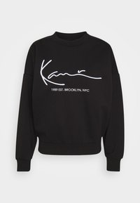 Sudadera negra de manga larga con cuello redondo. Presenta texto bordado en blanco que dice "Kamn" y "1989 EST. BROOKLYN, NYC." Textura suave.