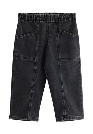Ung dreng med krøllet hår iført en hvid t-shirt, sorte løse jeans med store lommer og hvide sneakers, står med hænderne i lommerne.