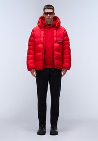 Chaqueta de plumas roja con capucha, puños elásticos y cremallera frontal. Lleva puesta una sudadera roja por debajo, combinada con pantalones negros y zapatos negros.