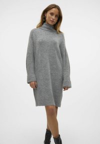 Abito maglione grigio con collo alto, maniche lunghe a costine e vestibilità oversize. Tessuto lavorato a maglia, lunghezza a metà coscia, abbinato a stivali neri al ginocchio.