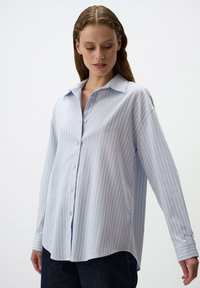 Camicia button-up azzurro chiaro con righe verticali bianche, maniche lunghe e colletto classico. Realizzata in tessuto morbido, presenta una vestibilità rilassata.