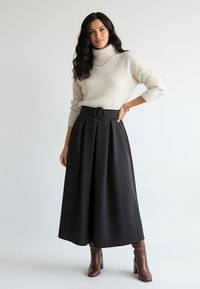 Kremowy, prążkowany sweter z golfem w zestawieniu z czarną, plisowaną spódnicą o wysokim stanie, ozdobioną okrągłą klamrą paska, oraz brązowymi botkami na obcasie.