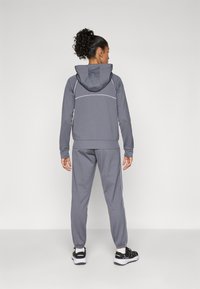 Gråidretts tracksuit med hettejakke og smale bukser med hvite detaljer og sidelister, laget av et glatt, fleksibelt materiale.
