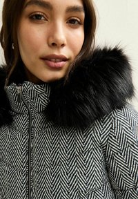 Kurtka puchowa w wzór chevron w czerni i bieli, z miękkim, czarnym kołnierzem z futra syntetycznego oraz metalowym zapięciem.