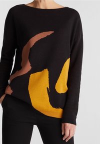 Svart tröja med abstrakta mönster i brunt och gult. Mjuk textur med vid halsringning och lös passform, kombinerad med svarta byxor.