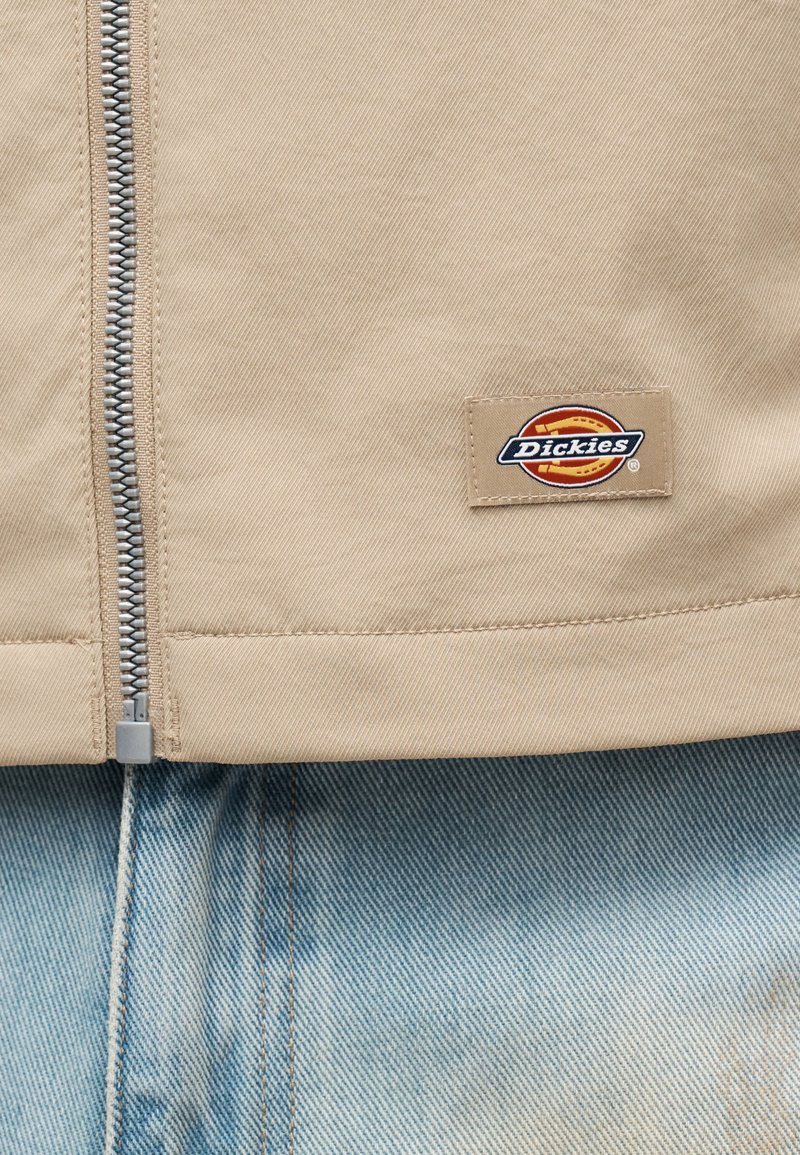Veste beige zippée avec un petit patch logo Dickies rectangulaire au-dessus d'un jean en denim bleu clair.