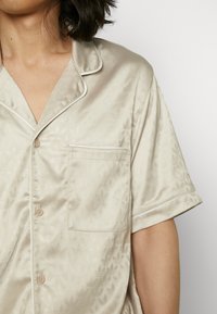 Ljust beige satintopp för pyjamas med kort ärm och en ficka, med ett subtilt geometriskt mönster och kontrasterande vit kant längs kragen.