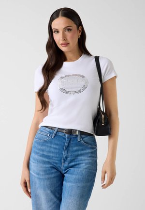 Donna con i capelli lunghi e scuri che indossa una t-shirt bianca a maniche corte con logo argentato, jeans blu, cintura nera e borsa nera a tracolla.