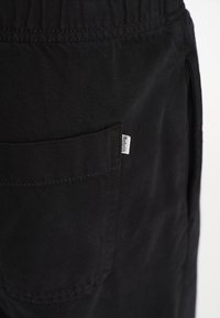 Sorte bomuldsbukser har et elastisk taljebånd, sidelommer og en baglomme med et mærke. Materialet har en glat tekstur.