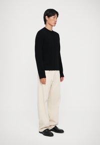 Sort strikketrøje med lange ærmer, parret med cremefarvede viddebukser og sorte loafers, på en neutral baggrund.