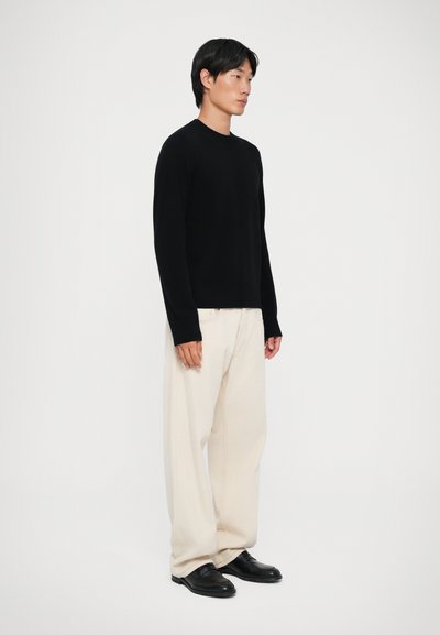 Maglione nero lavorato a maglia con maniche lunghe, abbinato a pantaloni larghi color crema e mocassini neri, su uno sfondo neutro.