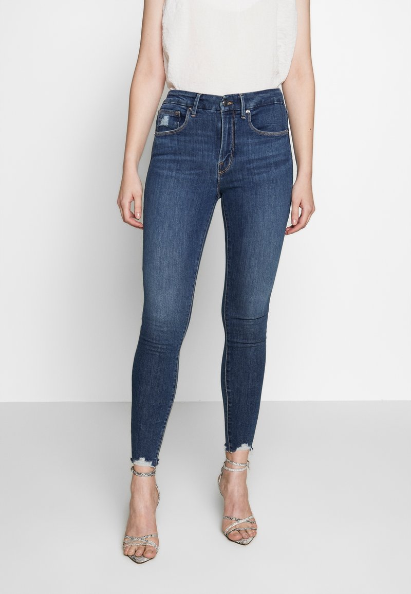 Tajta jeans i mörkblå denim med hög midja, med subtila slitningar och fransade kanter, bärs tillsammans med sandaler med remmar.