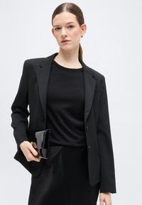 Γυναίκα με καστανά μαλλιά σε χαμηλή αλογοουρά, ντυμένη με μαύρο σακάκι, μαύρο τοπ και μαύρη φούστα, κρατώντας μια γυαλιστερή μαύρη τσάντα χειρός.