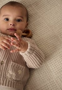 Hellbeige gestrickte Strickjacke mit einer Tasche und Sternen-Details, getragen von einem Säugling, auf einer strukturierten elfenbeinfarbenen Decke mit dezentem Muster.