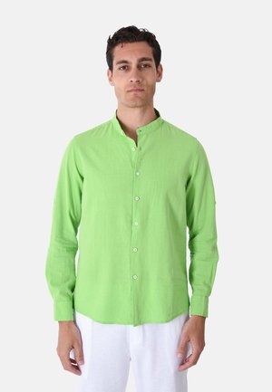 Uomo che indossa una camicia verde chiaro con colletto alla coreana abbottonata e pantaloni bianchi, in piedi davanti a uno sfondo bianco semplice.