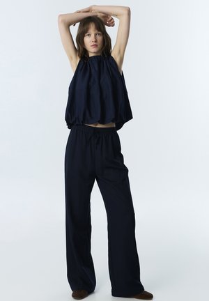 Jeune femme portant un haut noir ample sans manches et un pantalon à jambes larges, debout les bras levés au-dessus de la tête sur fond blanc.