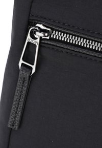 Détail d'un sac en tissu noir avec une fermeture éclair métallique et un tirette en cuir texturé, affichant le logo de la marque "JOOP!" embossé sur l'étiquette de la fermeture.