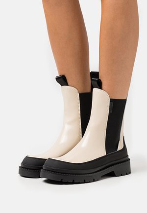 Beige en zwarte enkelboots met een glanzende afwerking, gestructureerde elastische zijkanten en een dikke, zwarte rubberen zool. Voorzien van trekbanden voor gemakkelijk aan- en uittrekken.