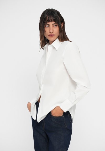 Camicia bianca a maniche lunghe con colletto classico, tessuto morbido e vestibilità rilassata, abbinata a pantaloni in denim scuro.