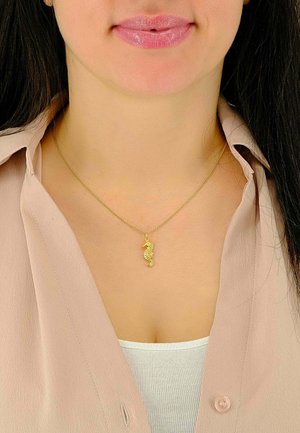 Pendentif en forme d'hippocampe doré sur une chaîne délicate, porté avec une chemise rose clair. Le pendentif présente des détails complexes et se détache sur un fond neutre.