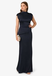 Žena v bezrukávových námořnických večerních šatech držící malou stříbrnou kabelku, nosící visací náušnice a zlaté sandály na otevřenou špičku, pózuje na bílém pozadí.