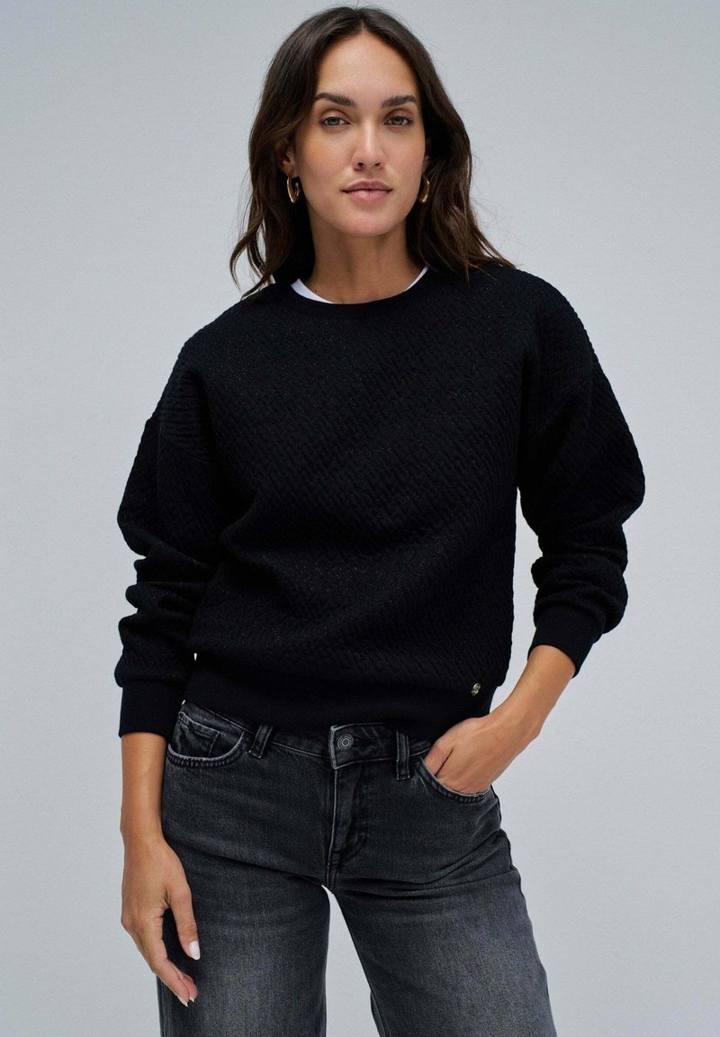 Černé strukturované mikina s kulatým výstřihem, spadlými rameny a žebrovanými manžetami. Přiřazeno k tmavě šedým džínům. Poznámka: jemný detail loga.