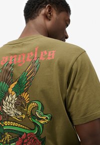Camiseta de color verde oliva con un gráfico colorido con un águila, una serpiente y una rosa en la parte posterior, estampado texturizado y texto estilizado "angeles".