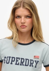 Hellblaues Baumwoll-T-Shirt mit marineblauem Rand, mit fettem "SUPERDRY"-Schriftzug und einem kleinen USA-Flaggen-Patch auf der linken Brust.