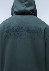 Felpa con cappuccio verde scuro caratterizzata da una stampa nera testurizzata e un motivo crepato, con etichetta "NAPAPIJRI geographic" sul retro.