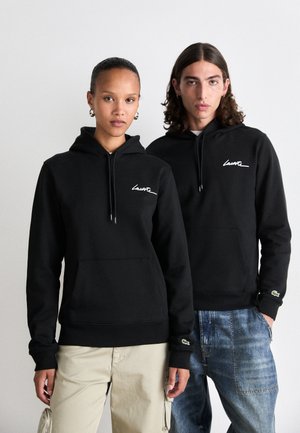 Two people wearing black hoodies with white Lacoste script logo, standing side by side against a plain light background.
