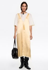 Lunghezza, abito di raso giallo chiaro con scollatura a V profonda, dettaglio con legatura davanti, bordi in pizzo, indossato sopra una maglietta bianca, abbinato a stivali neri e borsa.