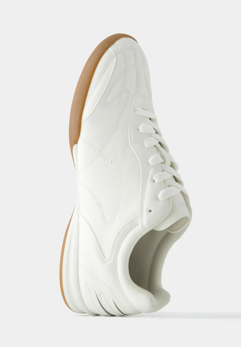 Białe sneakersy z teksturowaną skórzaną cholewką, sznurowanym zapięciem oraz brązową gumową podeszwą. Posiadają subtelne przeszycia i zakrzywioną sylwetkę.
