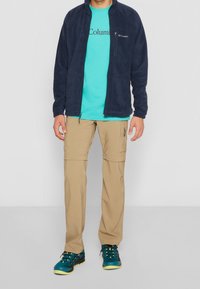 Giacca di pile blu navy, t-shirt turchese con logo, pantaloni cargo color khaki, scarpe da ginnastica blu e gialle, in piedi su uno sfondo neutro.